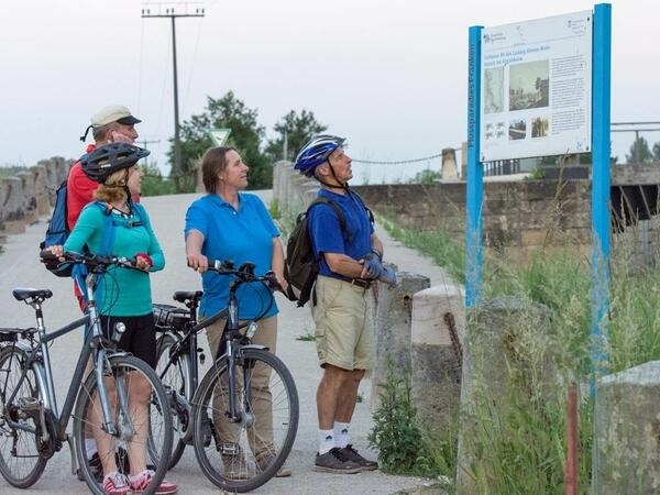 Vier Radfahrende informieren sich anhand einer Info-Tafel am Wegesrand.